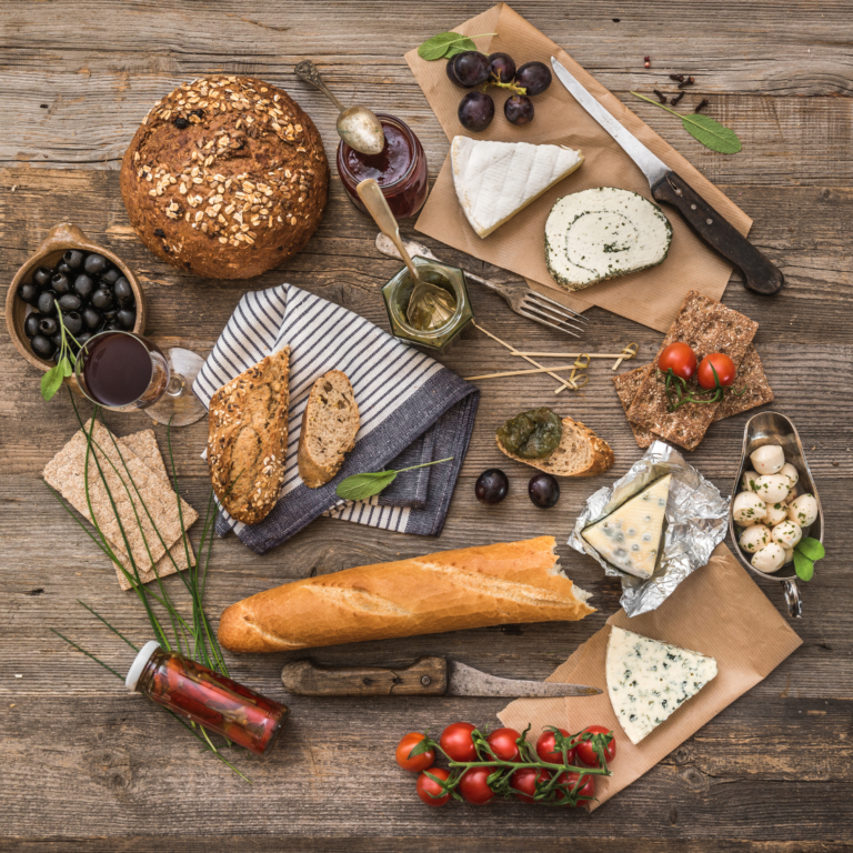 Food tours in France A rustic French food spread featuring artisanal cheeses, fresh baguette, olives, cherry tomatoes, grapes, and a glass of red wine on a wooden table. Perfect for a traditional French food tour experience.